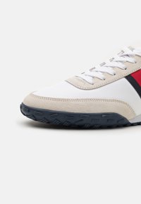 Lenkkari, jossa on valkoinen verkko-yläosa, beige mokkainen korostus, punaiset ja laivastonsiniset raidat sekä teksturoitu musta kumipohja.