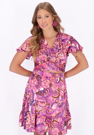 Fiatal nő hosszú, hullámos hajjal, élénk rózsaszín és lila virágmintás átvetős ruhában és hozzáillő fülbevalóval, mosolyogva, kezeit csípőjén tartva.