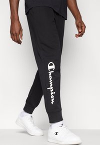 Czarne spodnie dresowe z białym logo "Champion" nadrukowanym po lewej stronie, charakteryzujące się wygodnym krojem i elastycznymi mankietami przy kostkach.