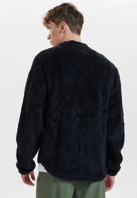 Svart fleecepullover med texturerad yta, långa ärmar och avslappnad passform. Har en rund halsringning och öppen rygg.