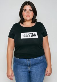 Młoda kobieta z ciemnymi, sięgającymi ramion włosami, ubrana w czarny t-shirt z napisem "BIG STAR" i niebieskie dżinsy, na jednolitym tle.