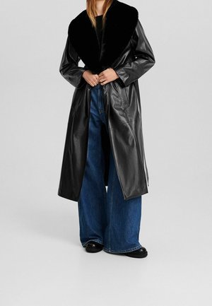 Person iført en lang sort læderjakke med stor pelskrave, brede blå jeans og sorte sko, stående mod en ensfarvet baggrund.