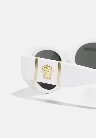 Weiße Sonnenbrille mit schwarzen Gläsern. Akzentuiert mit einer goldenen Klemme, die ein dekoratives Emblem trägt. Dicke Rahmen mit einem schlanken, modernen Design.