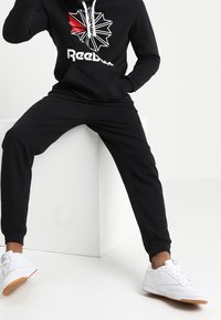 Black hoodie with a red and white graphic logo, paired with black tapered joggers and white sneakers with gum soles.