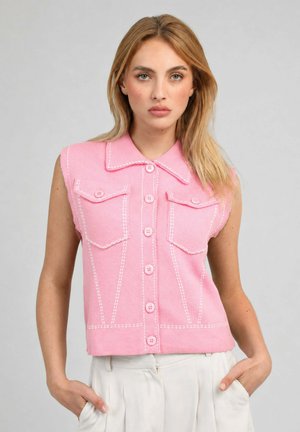 Femme blonde portant un gilet rose sans manches boutonné avec des coutures blanches et un pantalon blanc, debout les mains dans les poches devant un fond gris.