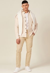 Camisa de botón color beige claro con botones oscuros, camiseta blanca debajo, suéter de punto crema caído sobre los hombros, pantalones color beige claro, zapatillas blancas.