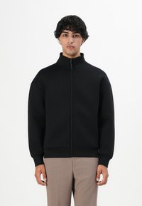 Chaqueta negra con cremallera y cuello alto, de mangas largas y puños y dobladillo acanalados. Textura suave, diseño minimalista. Lleva puesta sobre pantalones beige.