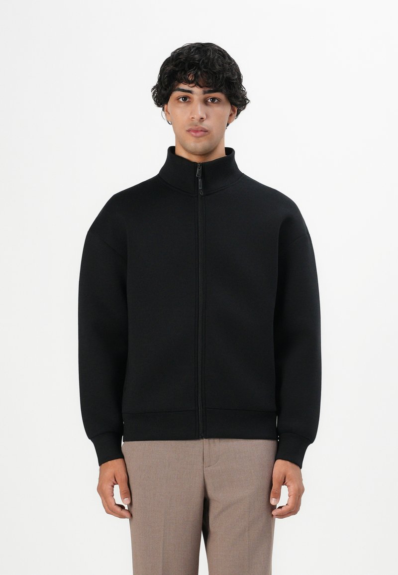 Chaqueta negra con cremallera y cuello alto, de mangas largas y puños y dobladillo acanalados. Textura suave, diseño minimalista. Lleva puesta sobre pantalones beige.