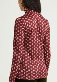 Top a maniche lunghe color granata con motivo geometrico ripetuto in beige e rosa. Tessuto morbido e liscio con silhouette aderente e collo alto.