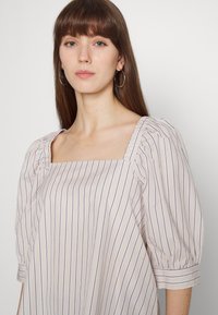 Blusa bege clara com riscas verticais azuis. Apresenta um decote quadrado, mangas bufantes e um corte solto. Textura de tecido suave.