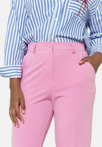 Roza krojena hlače z gladko teksturo, opremljene z gumbom in stranskimi žepi. V kombinaciji z modro in belo prugasto srajco.