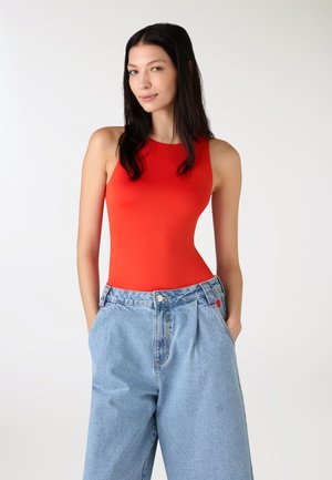 Donna con lunghi capelli scuri che indossa una maglietta rossa senza maniche e jeans a vita alta, gamba larga, di colore azzurro chiaro, con le mani nelle tasche.