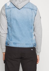 Chaleco de denim azul claro con una sudadera gris debajo. Presenta acentos desgastados y bolsillos traseros. Correas laterales ajustables. Pantalones negros visibles.