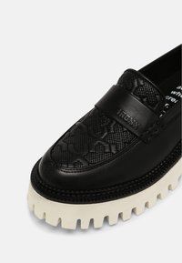 Mocassin en cuir noir avec un dessus texturé, nom de la marque embossé sur la sangle et semelle en caoutchouc blanche épaisse et nervurée.