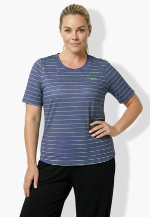 Frau mit blondem Pferdeschwanz, die ein blau-grün gestreiftes kurzärmeliges Hemd und schwarze Hosen trägt, steht mit einer Hand in der Hüfte.