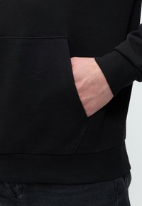Hand in die Fronttasche eines schwarzen Langarm-Hoodies gesteckt, getragen mit schwarzen Hosen.