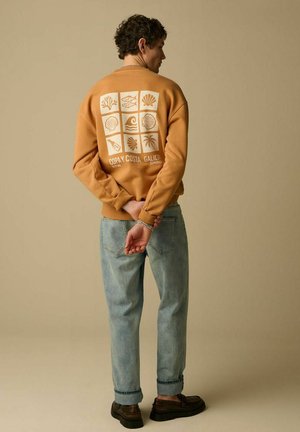 Homme en jean bleu clair et sweat-shirt beige avec des motifs et du texte sur le thème de la mer, debout avec les mains derrière le dos sur un fond beige.