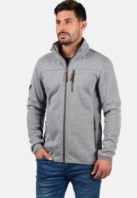 Veste zippée grise en tissu texturé, avec des accents en cuir marron sur la fermeture éclair et les épaules, dotée d'un col montant et de poches latérales.