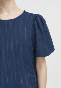 Marineblauwe top met pofmouwen, gemaakt van gestructureerde stof met subtiele verticale strepen en een ronde halslijn. Eenvoudig, ontspannen ontwerp.