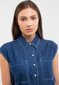 Tamsiai mėlynas džinsinis marškinis su apykakle, trumpomis rankovėmis, baltais siūlėmis ir dviem krūtinės kišenėmis su mygtukais.