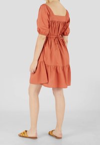 Robe couleur rouille à manches bouffantes, décolleté carré, détails froncés à la taille et ourlet à volants. Associée à des sandales jaunes à enfiler.