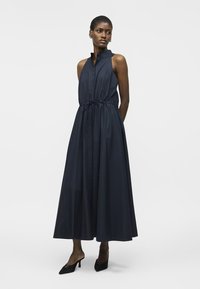 Femeie purtând o rochie midi bleumarin fără mâneci, cu guler înalt, talie strânsă și pantofi cu toc ascuțit negri, stând în fața unui fundal simplu.