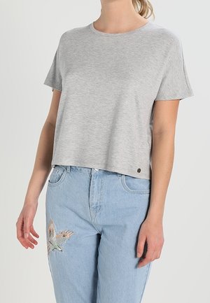 Femme portant un T-shirt court à manches courtes uni gris clair et un jean bleu clair avec une broderie d’oiseau sur la partie supérieure de la cuisse.