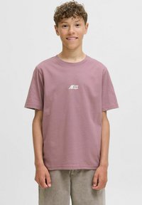 Jonge persoon met krullend haar glimlacht, draagt een lichtpaarse t-shirt met korte mouwen en een wit logo, staande voor een effen achtergrond.