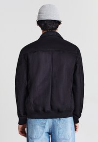 Persona che indossa una giacca nera, jeans azzurri chiari e un berretto grigio chiaro, girato di spalle su uno sfondo chiaro e semplice.