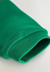 Gros plan sur un poignet de manche en tissu tricoté vert avec une texture côtelée sur fond clair.