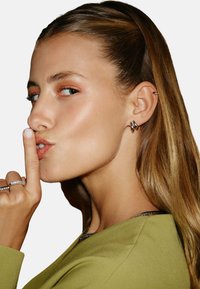 Modell i en grön topp med ett silverkedja. Närbild i profil som visar långt hår, ringörhängen och finger mot läpparna i en tystnadsgest.