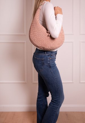 Frau trägt ein weißes Langarmshirt und blaue Jeans mit großer rosa gewebter Handtasche über der Schulter vor einem blassen Wandhintergrund.