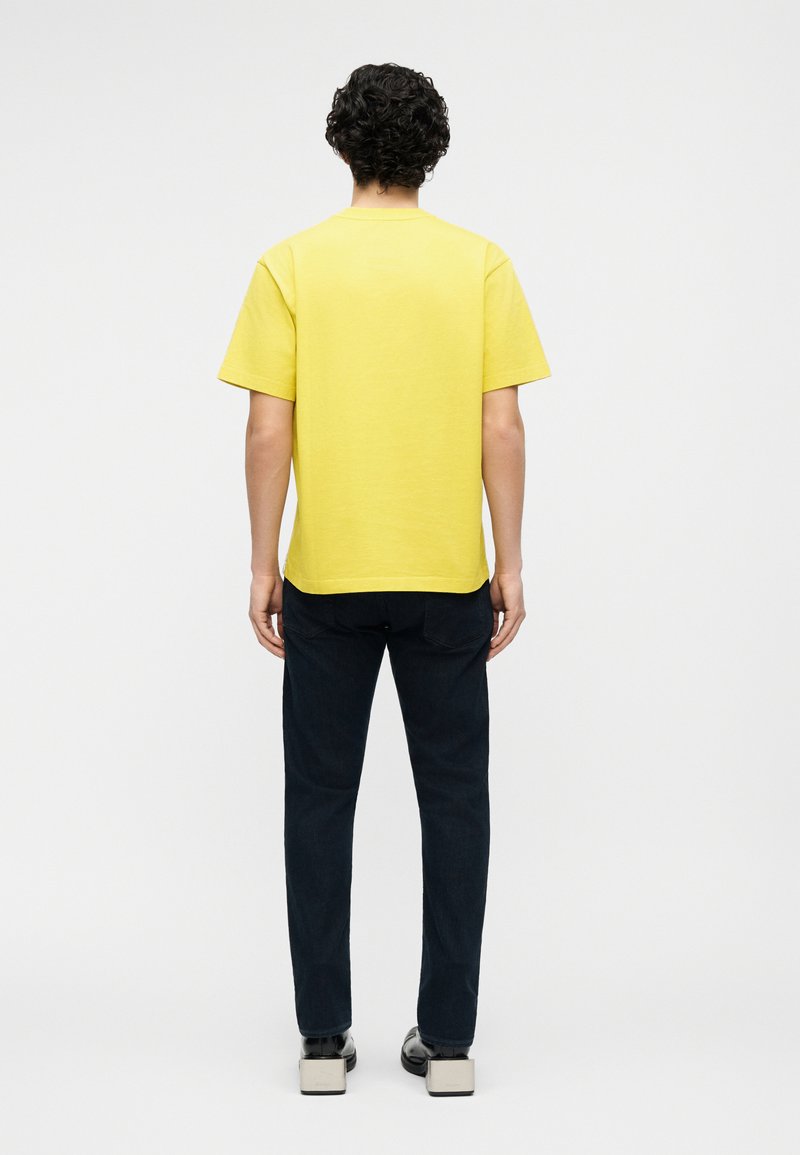 Uomo con capelli ricci che indossa una maglietta gialla a maniche corte semplice, pantaloni scuri e scarpe nere, in piedi di spalle su uno sfondo bianco.