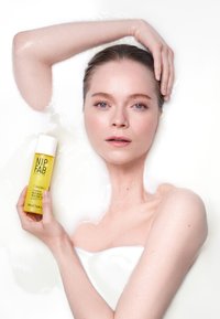 Brume hydratante NIP+FAB dans une bouteille jaune, avec un capuchon blanc ; tenue par une personne dans l'eau, mettant en valeur une peau lisse et un design épuré.