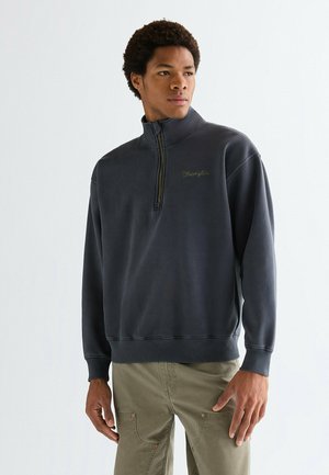Ung mand iført en mørkegrå zip-up sweatshirt med "Wrangler" broderi, kombineret med lyse olivengrønne cargobukser, stående mod en ensfarvet baggrund.