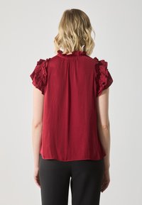 Rød bluse med volang korte ermer, høy hals og lett stoff. Bakdelen er litt lengre med en myk, flytende fall.