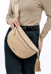 Tan leren schoudertas met een gebogen ontwerp, ritsluiting en een franjeraccent. Draagt over een donkere outfit met een geweven riem.