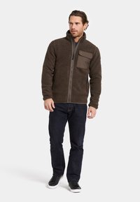 Bruine fleece jasje met een ritssluiting aan de voorkant, hoge kraag en een grote borstzak van een contrasterend materiaal, gecombineerd met donkere spijkerbroek en zwarte sneakers.
