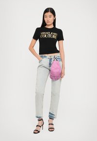 Melnas īsās piedurkņu T-krekls ar zelta "VERSACE JEANS COUTURE" logo, gaiši zilas augstās jostas vietas džinses, rozā cepures soma, melni papēži.