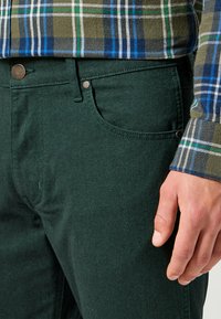Donkergroene katoenen broeken met gestructureerde stof, vijf zakken en metalen accenten. Gedragen met een blauw-groene geblokte flanellen shirt.