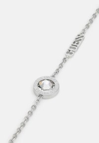 Silberarmband mit einem runden Anhänger, der einen klaren Kristall in der Mitte hat, graviert mit "GUESS", und Kettendesign.