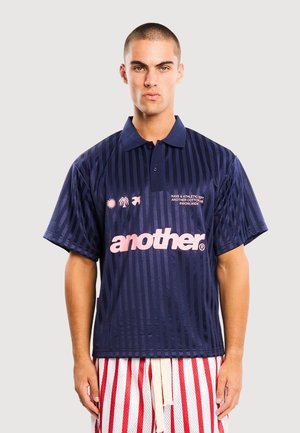 Junger Mann mit kurzen Haaren, der ein marineblau gestreiftes Polohemd mit pinkem Schriftzug „another“ und rot-weiß gestreifte Shorts trägt.