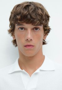 Polo bianco in tessuto texturizzato con colletto. Il soggetto ha capelli corti e ondulati di colore marrone e uno sfondo neutro.
