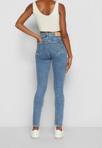 Kvinna i ljusblå tajta jeans, vit cropped tank top och vita sneakers, stående med ena handen i bakfickan mot en enkel bakgrund.