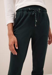 Tmavě zelené kalhoty s elastickým pasem a šňůrkou, které mají boční kapsy a hladkou texturu látky. Design obsahuje stříbrné kovové akcenty.