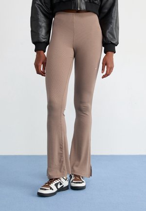 Osoba ubrana w wysokie, rozkloszowane leginsy w kolorze taupe, czarną krótką skórzaną kurtkę i brązowo-biało-czarne sneakersy stoi na niebieskiej podłodze.