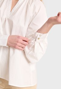 Weiße Baumwollbluse mit V-Ausschnitt, voluminösen Ärmeln mit Knopfakzenten und lockerer Passform. Glatte Textur und leichtes Material.
