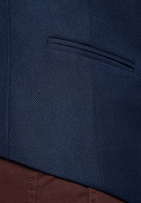 Granatowa, teksturowana tkanina marynarki o dopasowanym kroju z pojedynczą kieszenią na piersi, zestawiona z brązowymi spodniami, które są widoczne na dole.