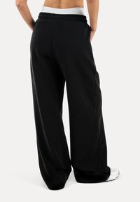 Osoba ubrana w luźne czarne spodnie z elastycznym pasem i białe trampki, pokazana od tyłu na jednolitym tle.