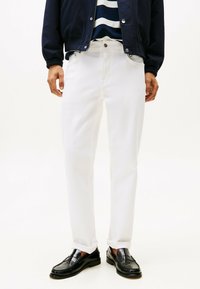 Jeans di denim bianchi con una vestibilità slim, dotati di cinque tasche, chiusura frontale con bottone e orli arrotolati, abbinati a mocassini neri.